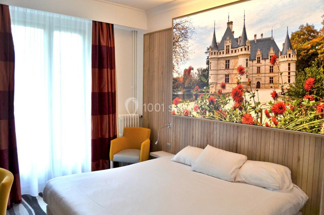 Chambre lumineuse avec lit double, fauteuils, grande fenêtre et tableau représentant un château entouré de fleurs.