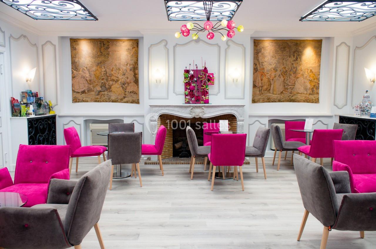 Salle lumineuse avec des fauteuils roses et gris, tables en verre, cheminée décorative et tapisseries murales.