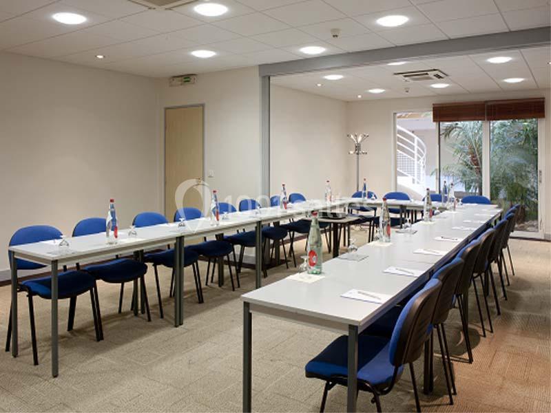 Salle de réunion lumineuse avec des tables disposées en U, des chaises bleues et des bouteilles d'eau sur les tables.