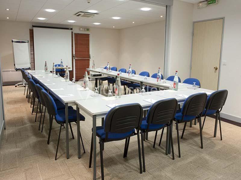 Salle de réunion lumineuse avec tables disposées en U, chaises bleues, tableau blanc et écran de projection.
