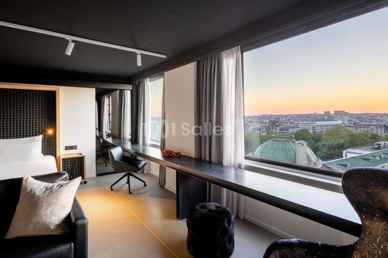 Chambre d'hôtel moderne avec lit, bureau, canapé et vue panoramique sur Bruxelles au coucher de soleil.