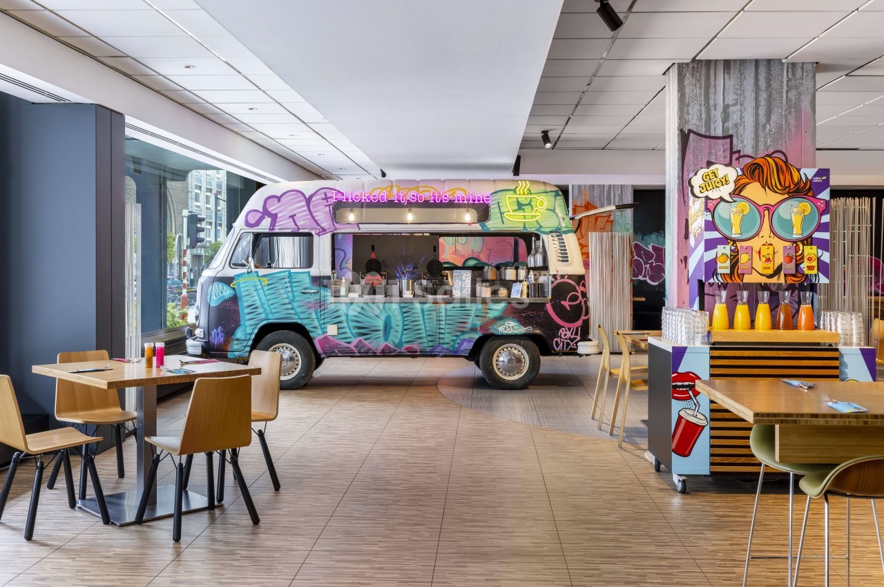 Salle d'événement avec food truck coloré customisé, tables en bois et chaises design, bar pop art en arrière-plan.