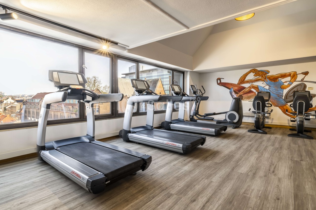 Salle de fitness équipée de tapis roulants, vélos elliptiques et art mural, baies vitrées vue sur la ville.