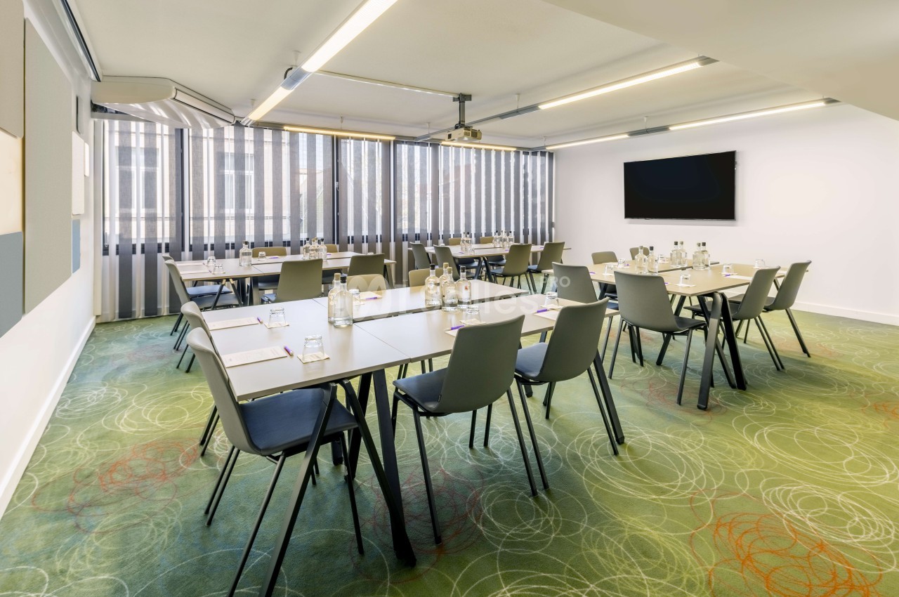 Salle de réunion modulable avec tables blanches, chaises vertes, baies vitrées et écran mural.
