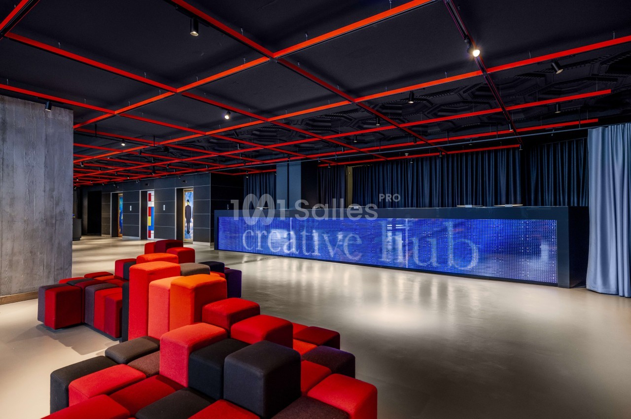 Salle moderne aux néons rouges au plafond, poufs rouge et noir, écran LED bleu, portes grises et rideaux bleus.