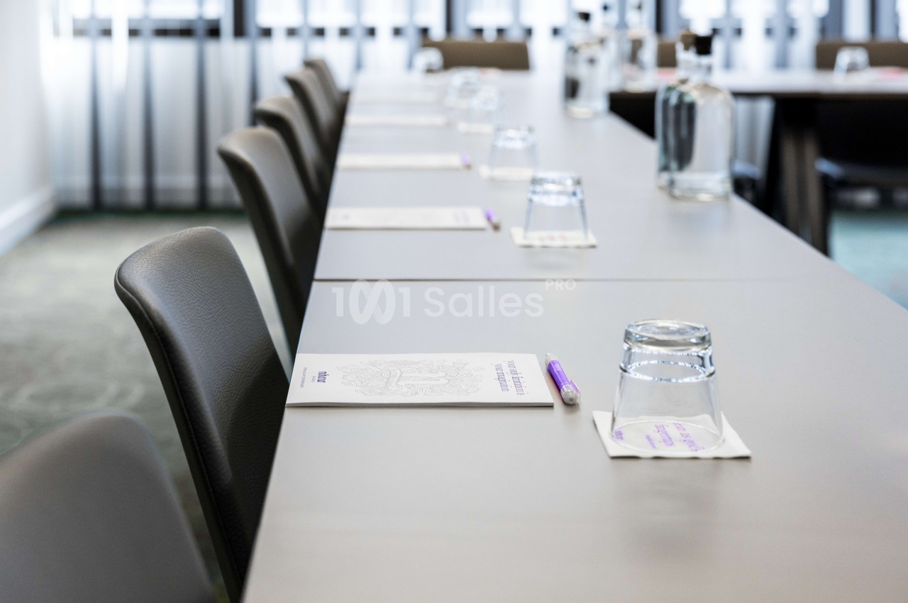 Salle de réunion avec table longue, chaises grises, verres d'eau et bloc-notes - lumière naturelle.