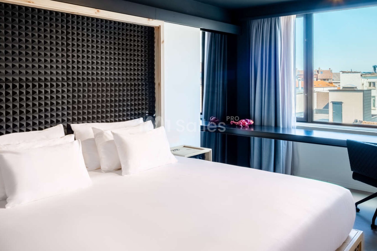 Chambre d'hôtel avec lit blanc, tête de lit texturée noire et vue urbaine par baie vitrée.