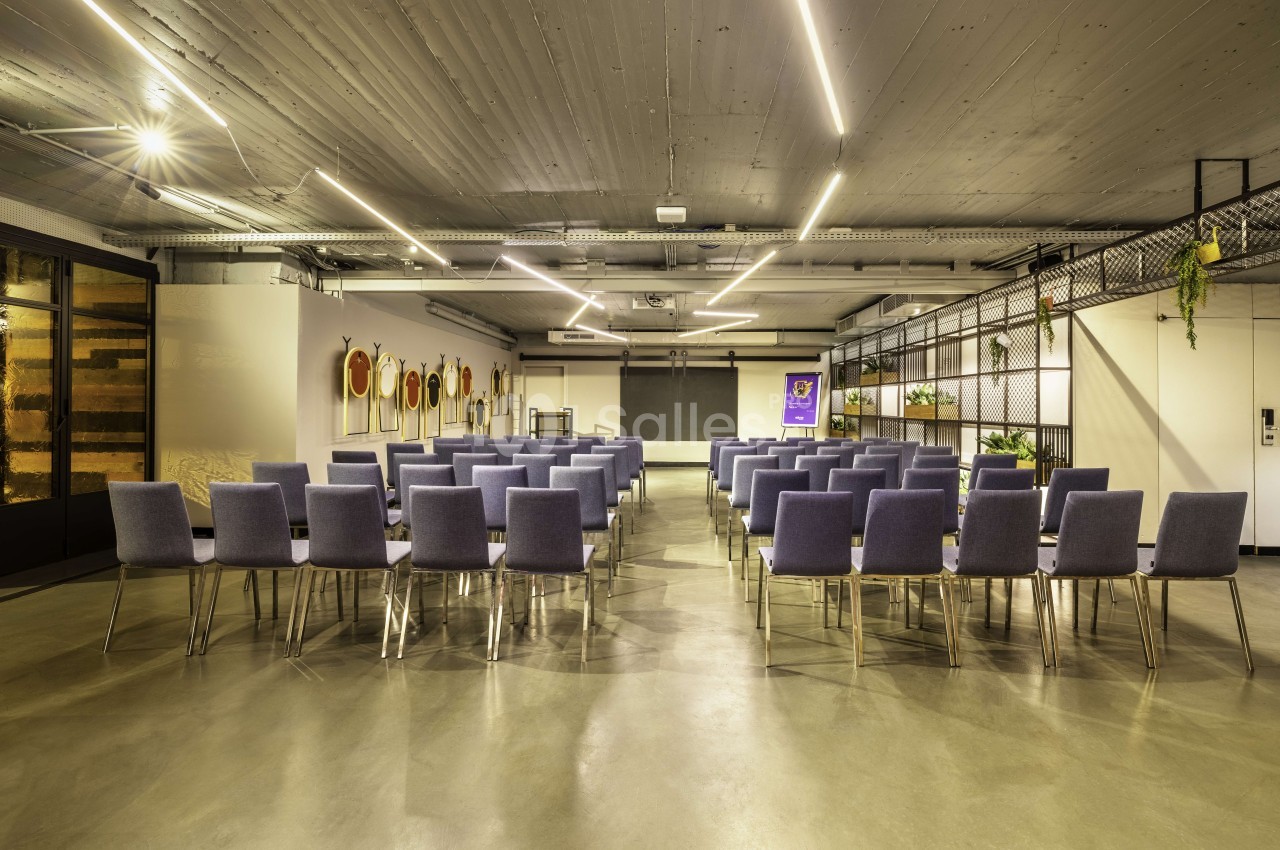 Salle de conférence avec chaises grises en rangées, sol béton, plafond industriel et éclairage LED linéaire.
