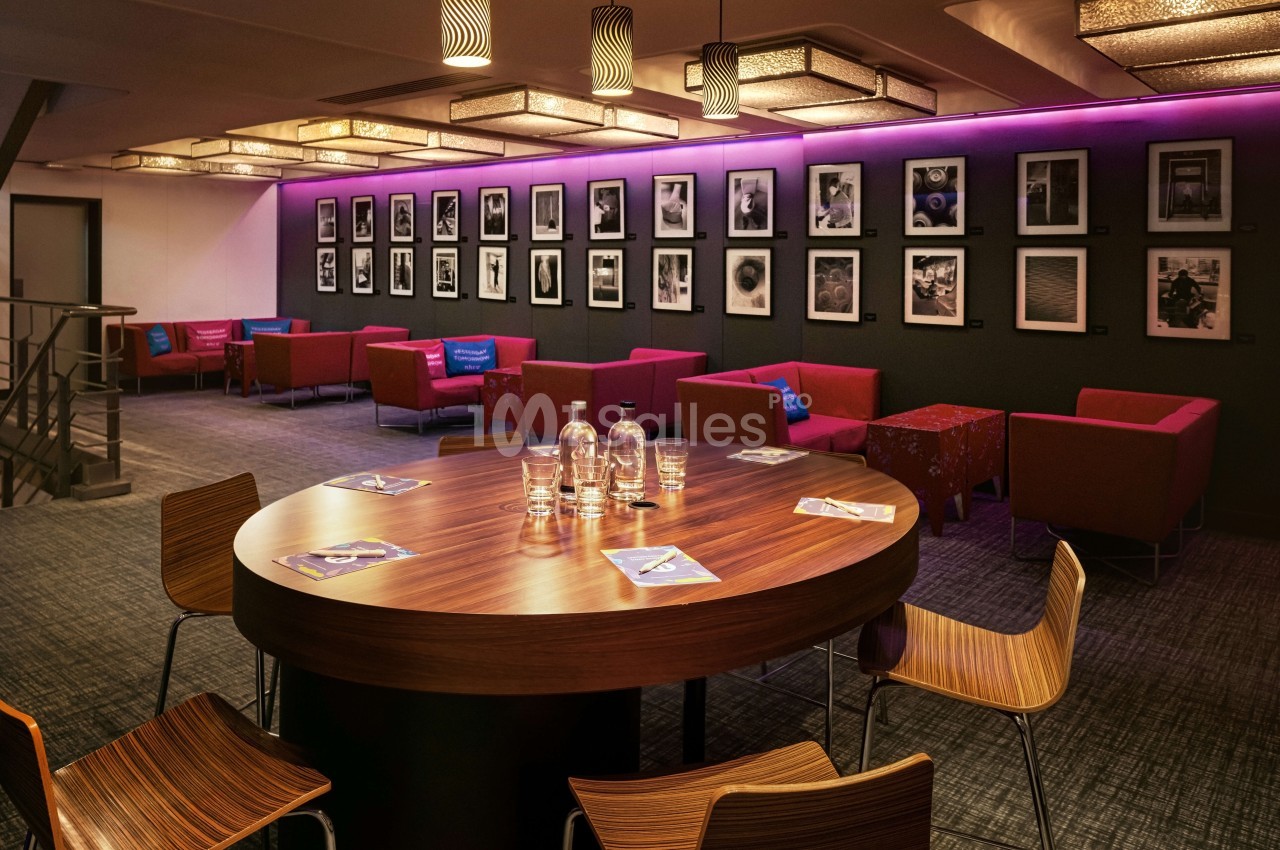 Salle de réunion avec table ovale en bois, chaises design, canapés rouges et mur violet orné de cadres photographiques.