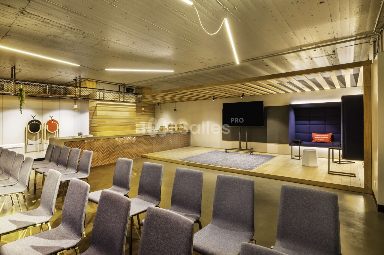 Salle de conférence moderne avec chaises grises, estrade surélevée, écrans et bar en arrière-plan - Nhow Brussels.