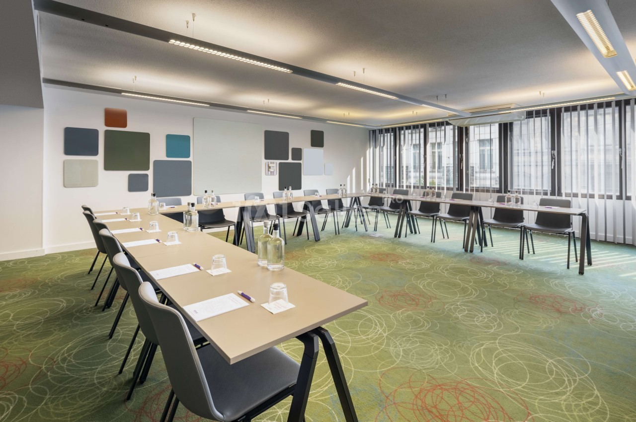 Salle de réunion avec tables rectangulaires dressées, chaises noires, murs blancs, panneaux colorés et grandes baies vitrées.