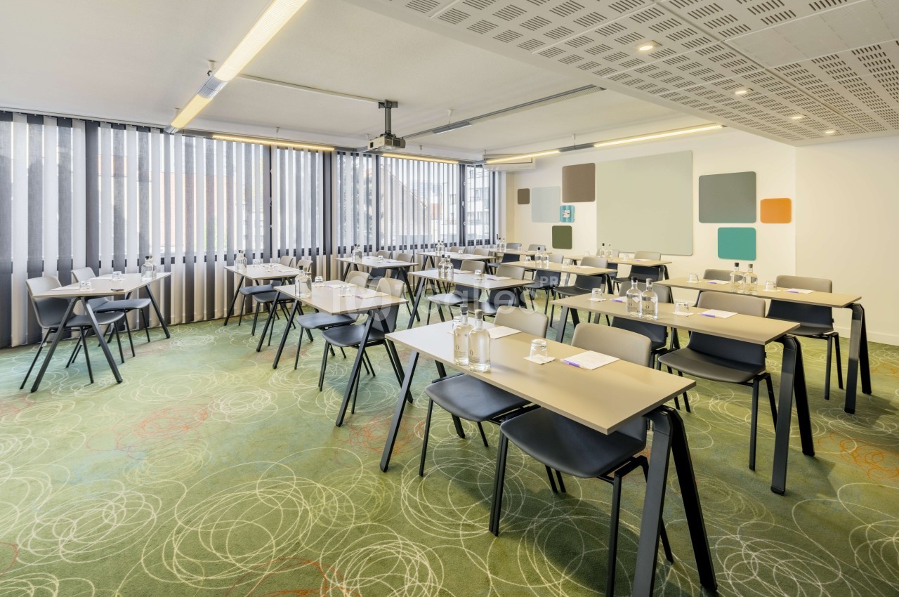 Salle de réunion dressée en rangées avec tables beiges, chaises noires, baies vitrées et sol motifs géométriques verts.