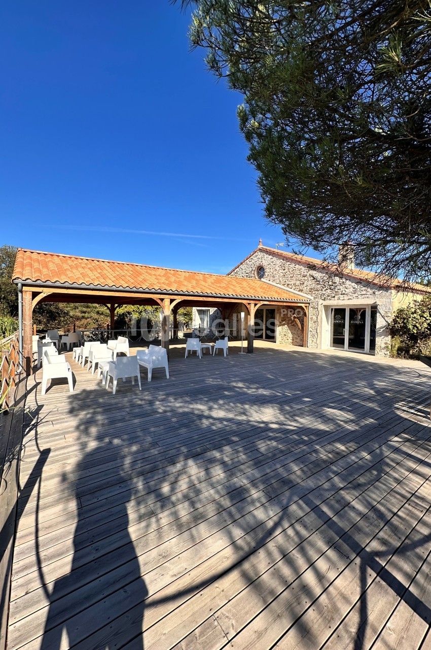 Terrasse en bois avec chaises blanches, pergola couverte et bâtisse en pierre sous ciel bleu dégagé.