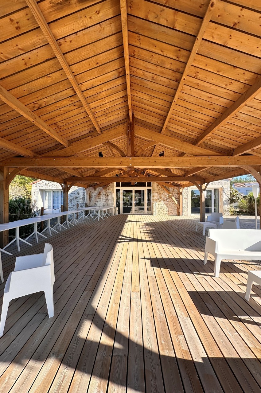 Terrasse couverte en bois sous structure en poutres apparentes, chaises blanches disposées, accès à bâtiments en pierre.