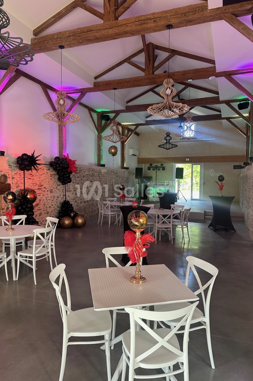 Salle aménagée avec poutres apparentes, tables rondes blanches, chaises blanches et décoration événementielle rouge et or sous éclairage rose et violet.