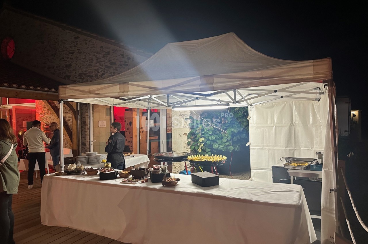 Espace de réception nocturne sous chapiteau blanc avec buffet dressé, convives et cuisines extérieures.