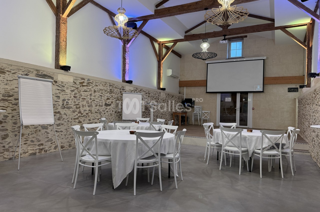 Salle intérieure avec poutres apparentes, murs en pierre, tables rondes dressées de nappes blanches et chaises blanches, éclairage bleu et vert.