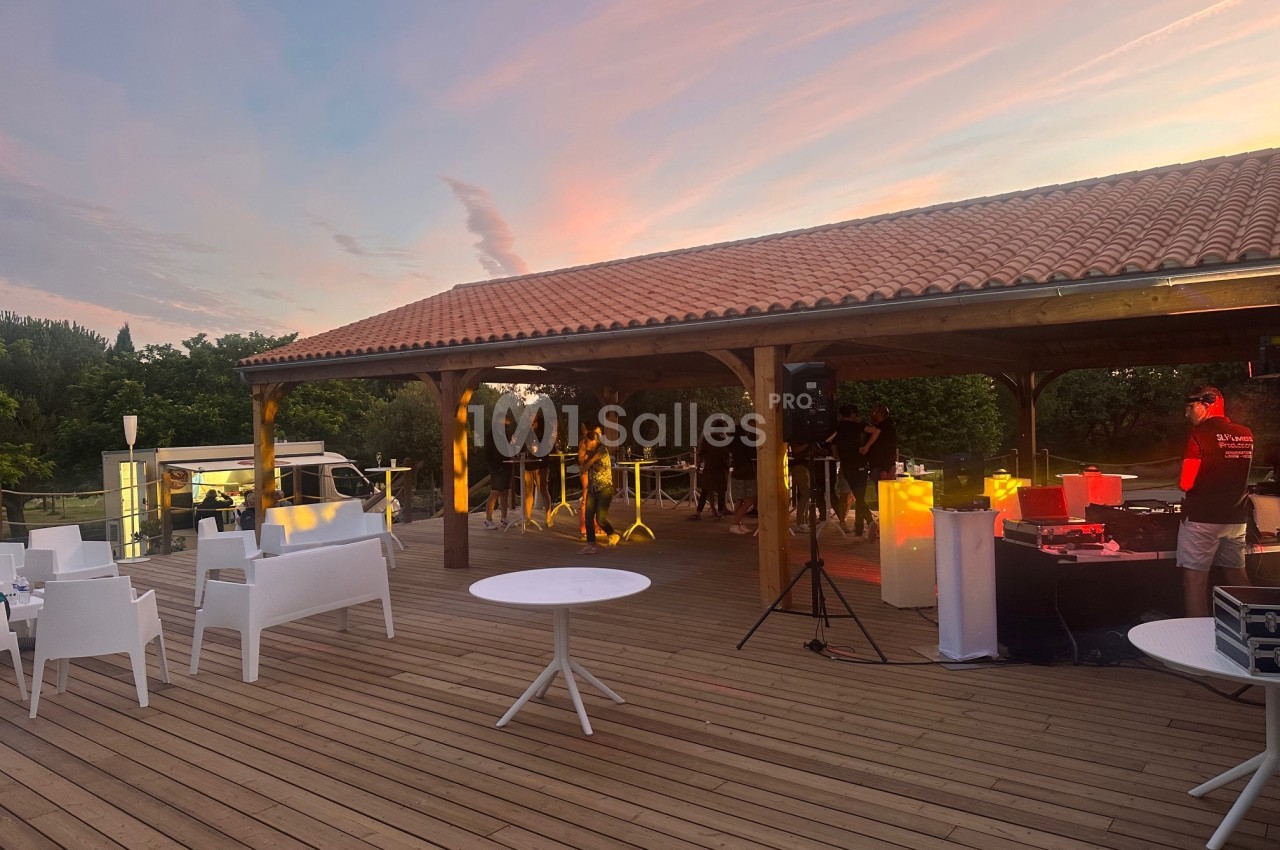 Terrasse couverte avec charpente bois, tables hautes blanches et mobilier, éclairage ambiant au coucher de soleil.