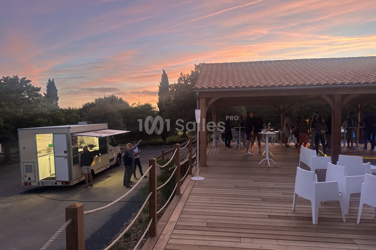 Terrasse en bois avec tables hautes, chaises blanches, food truck et abri couvert, coucher de soleil en arrière-plan.
