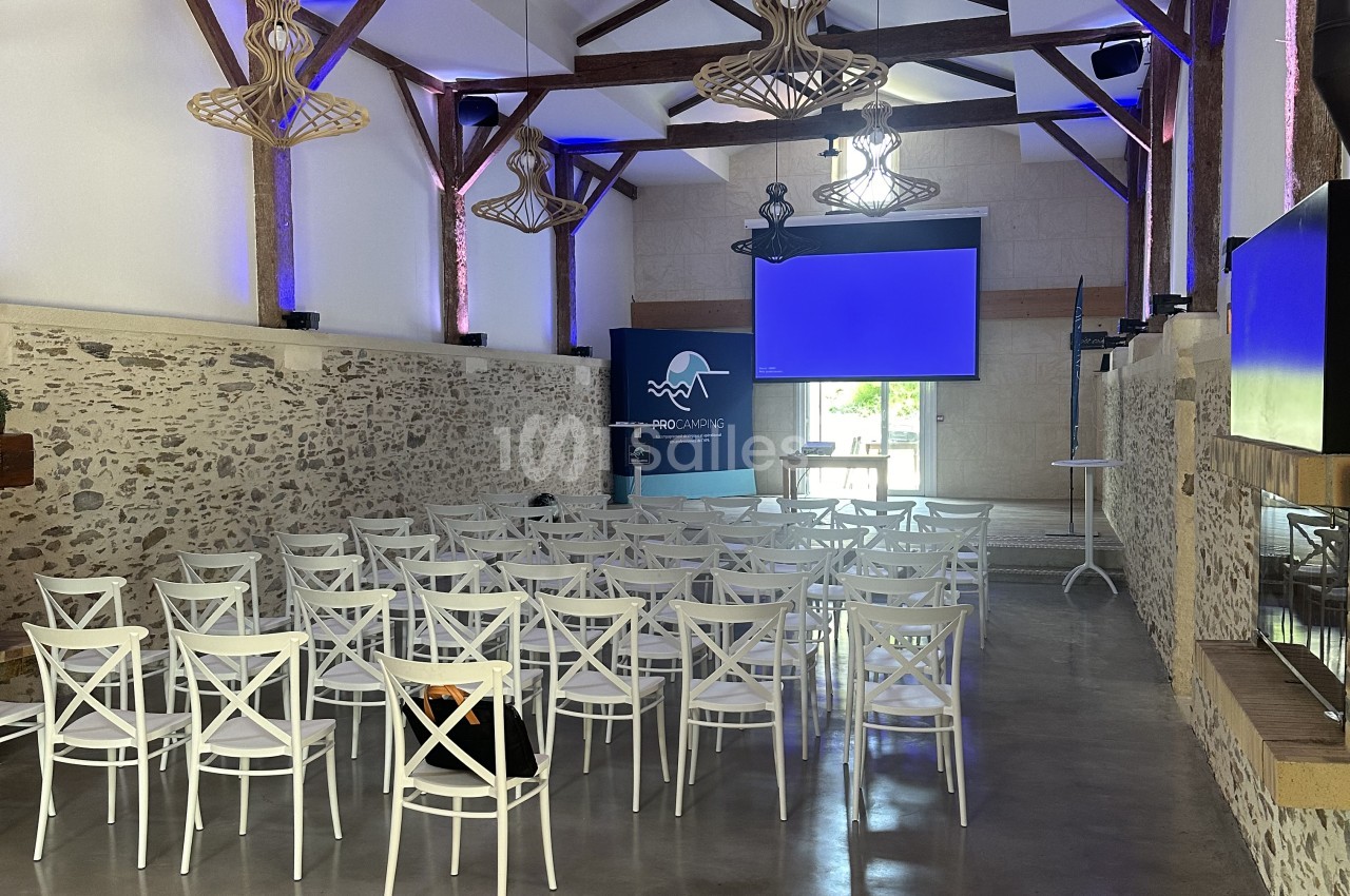 Salle avec poutres apparentes, chaises blanches alignées, murs en pierre, éclairage bleu et écran.