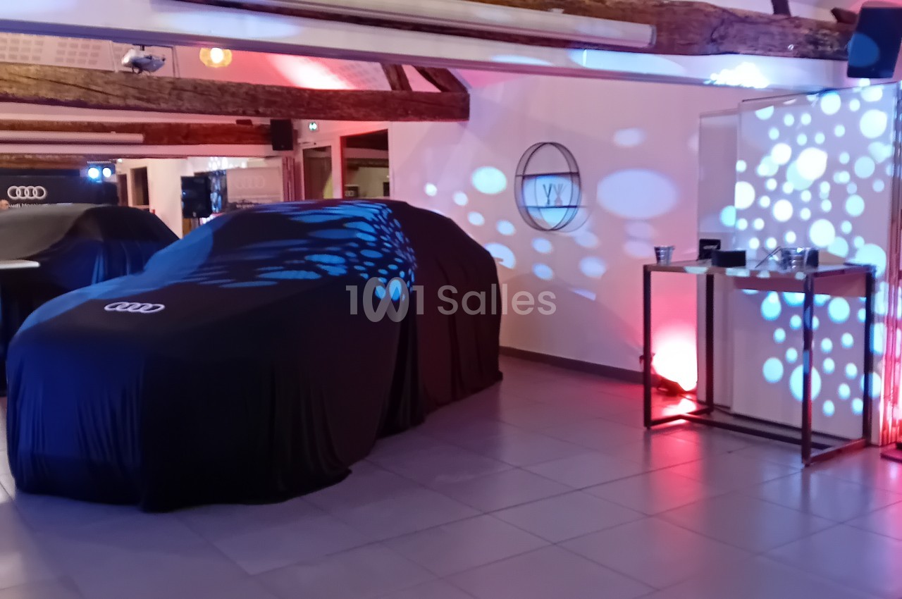 Salle d'exposition avec voiture Audi sous housse noire, poutres apparentes, éclairage rose et bleu, écrans lumineux.