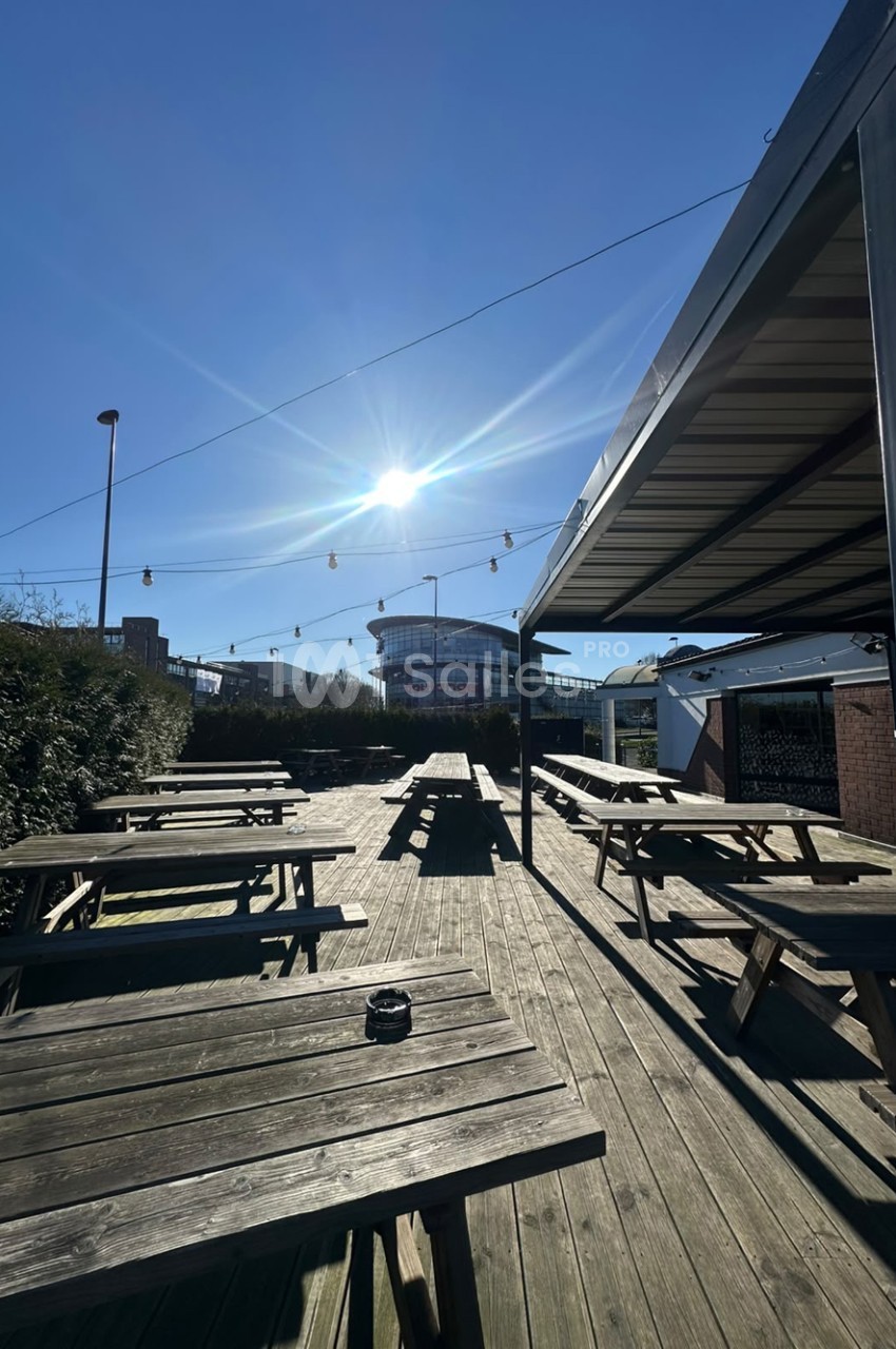 Terrasse en bois extérieure sous un ciel bleu, avec des tables et bancs de pique-nique alignés sous une pergola et des guirlandes lumineuses.