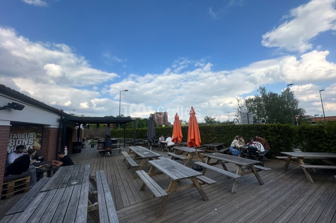 Terrasse en bois extérieure avec tables de pique-nique et parasols orange, quelques personnes attablées.