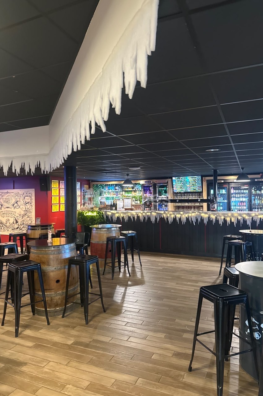 Intérieur d'un bar à l'ambiance sombre avec plafond noir, tables hautes en fûts de bois, tabourets noirs, comptoir décoré de guirlandes lumineuses blanches et étagères de bouteilles. Let me check length: roughly 170 characters, too long. Need ≤125.