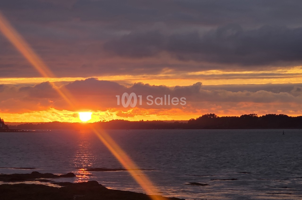 Coucher de soleil sur la mer avec rayons lumineux, forêt à l'horizon et rochers en premier plan.