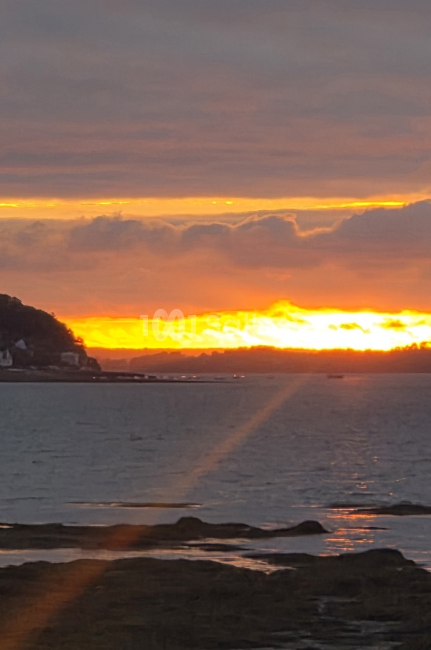 Coucher de soleil sur l'océan avec ciel orangé, nuages dorés et rochers en premier plan.