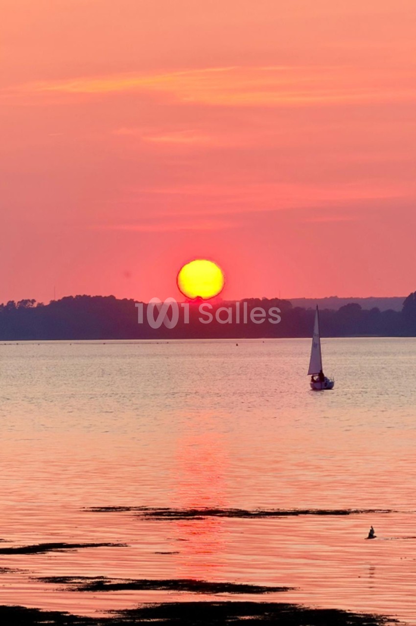 Coucher de soleil sur une baie avec voilier au mouillage et boisé en arrière-plan.