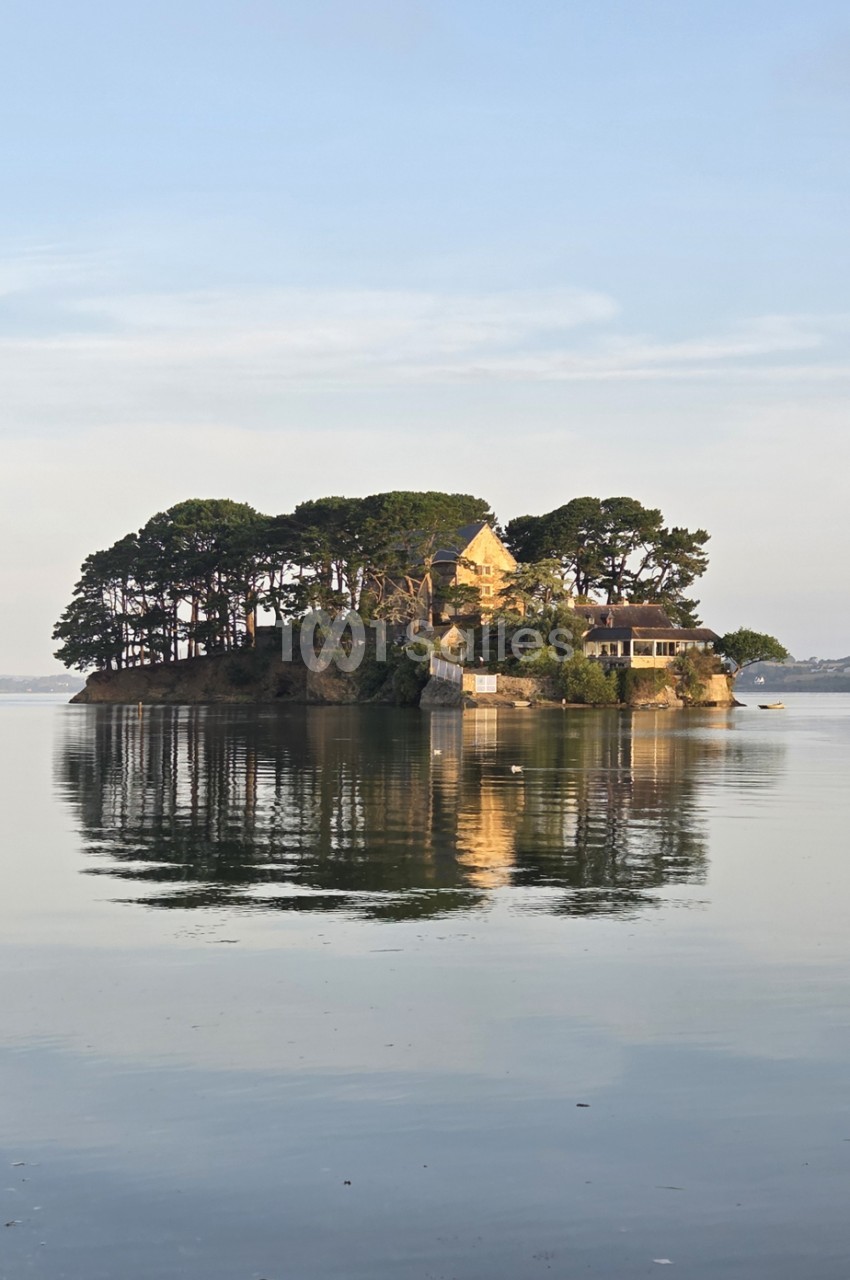 Petite île boisée avec maison de pierre jaune clair, entourée d'eau calme et reflétée.