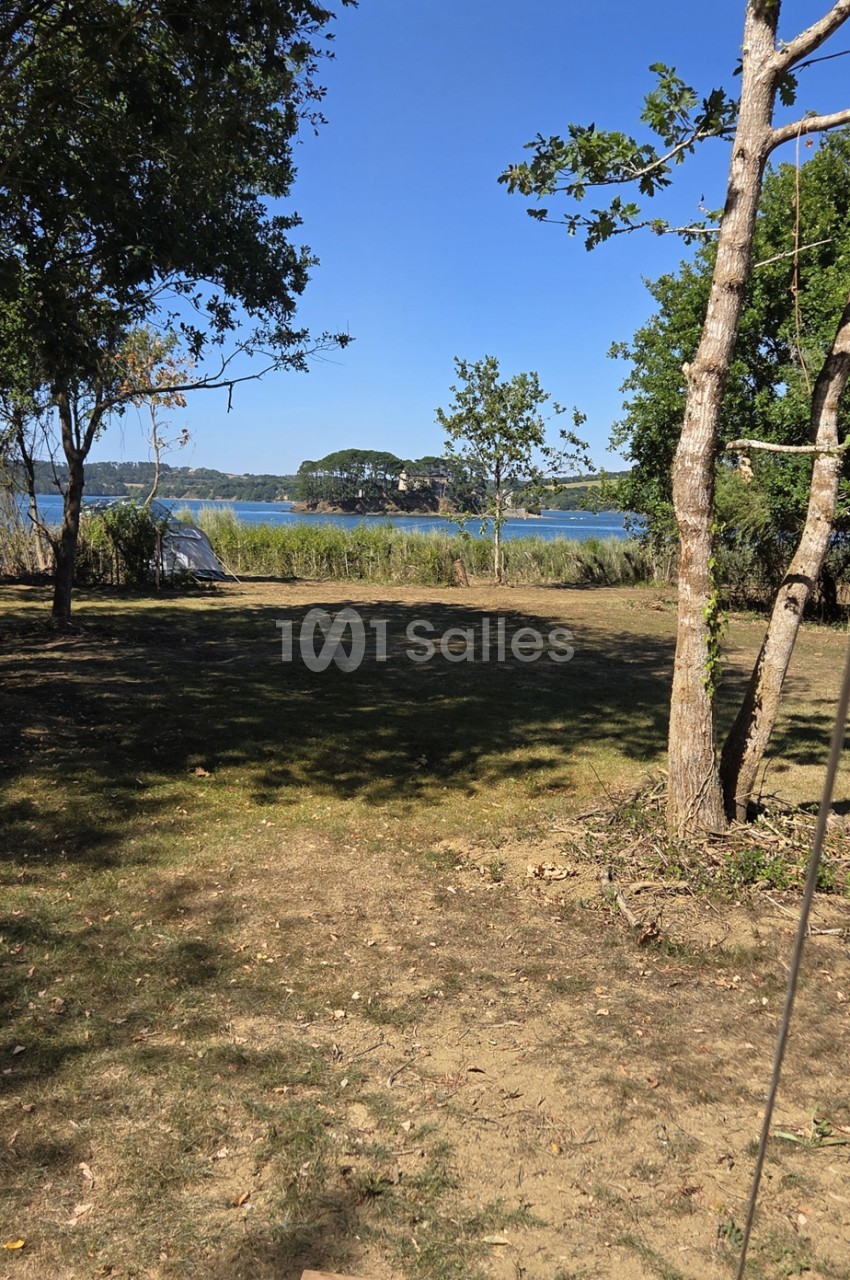 Espace extérieur herbeux ombragé par des arbres, vue sur lac calme avec rives boisées en arrière-plan.