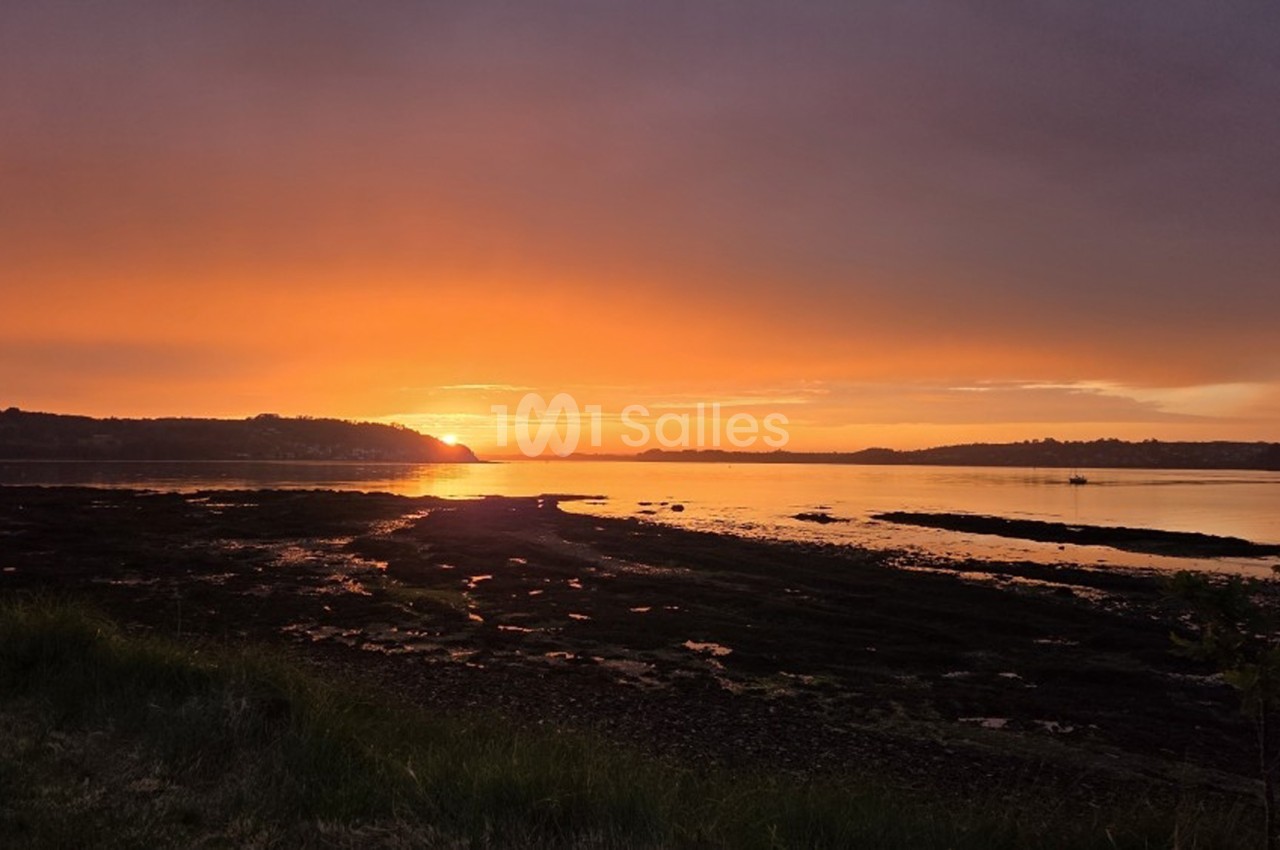 Coucher de soleil doré sur une baie avec côtes boisées et plage rocheuse au premier plan.