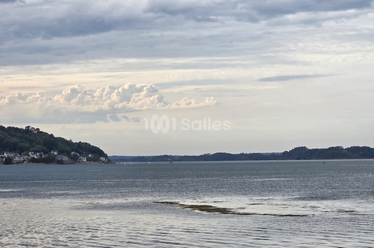 Baie côtière avec maisons sur les falaises boisées, ciel nuageux et mer calme en avant-plan.