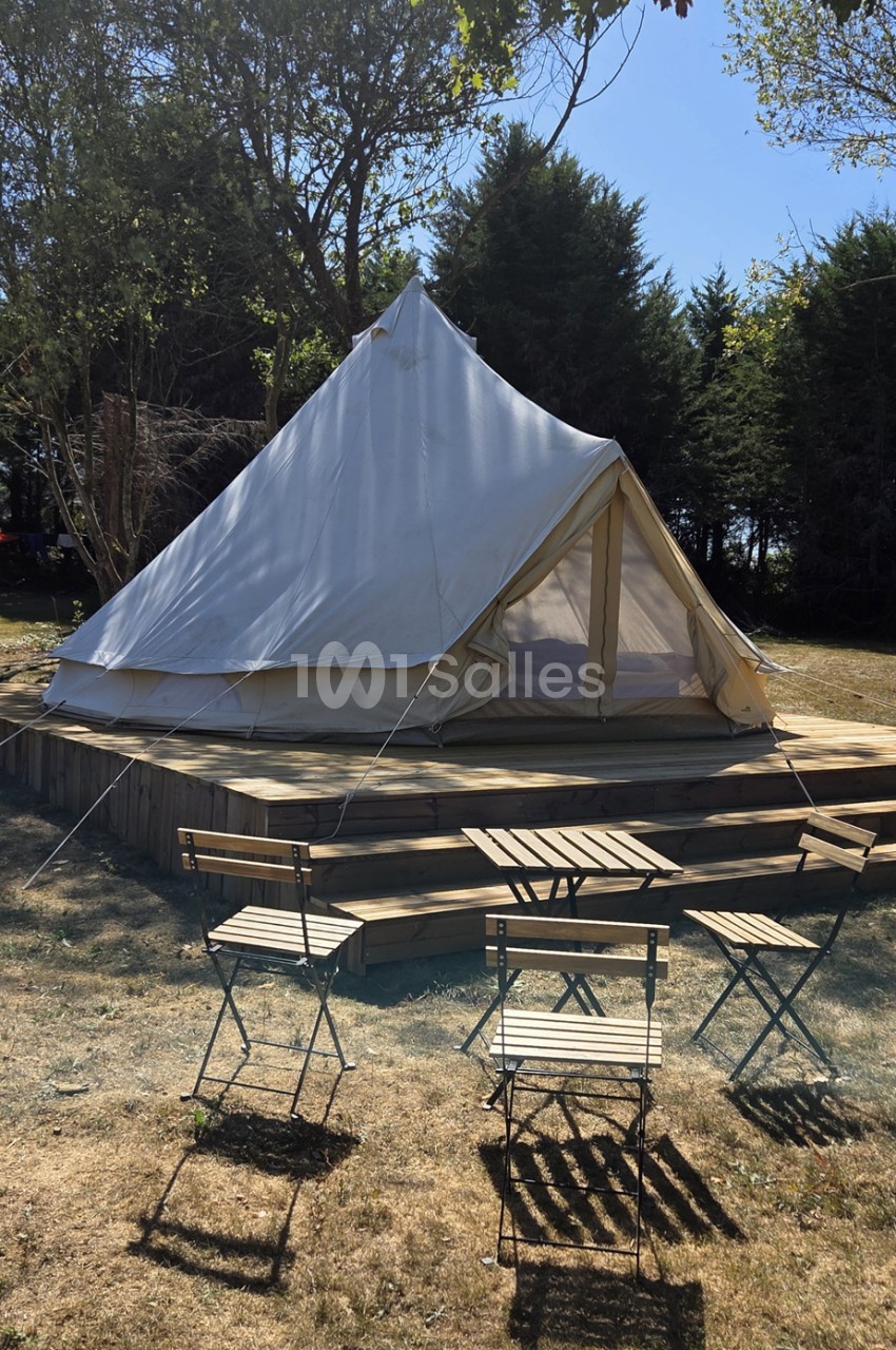 Tente glamping blanc et beige sur plateforme bois, entourée de chaises pliantes, en forêt.
