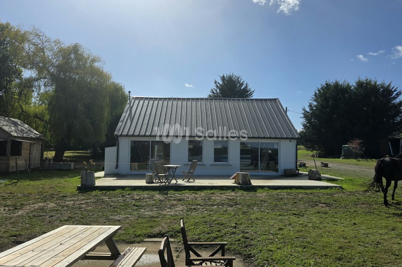 Petite maison blanche à toit métallique gris avec grandes baies vitrées, entourée d'herbe, d'arbres et d'une terrasse en béton avec table et chaises.