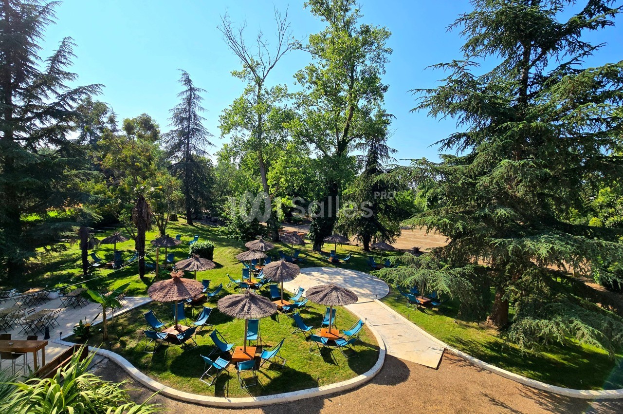 Parc arborisé avec espaces lounges ombragés - transats bleus et parasols en paille disposés sur pelouse sous arbres centenaires.