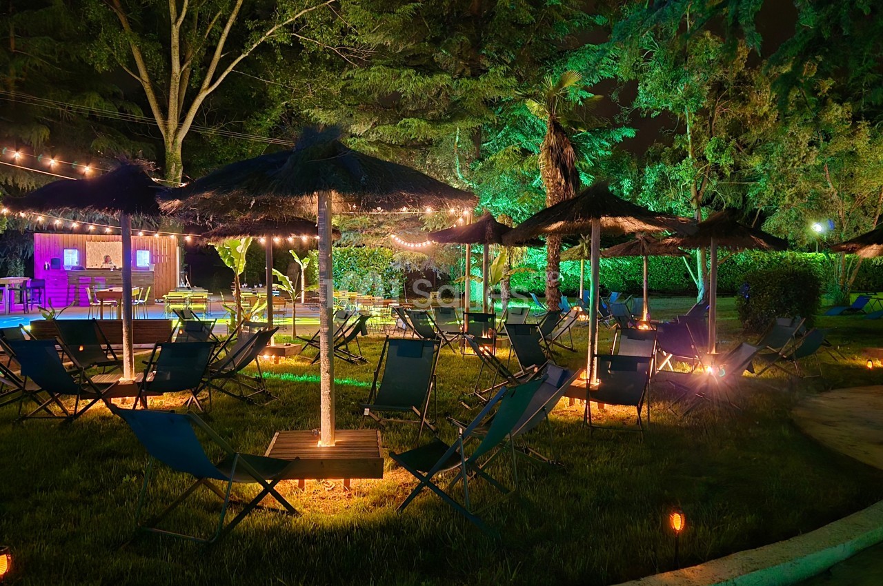 Espace extérieur de nuit avec chaises longues sous parasols, éclairage coloré vert et or, bar aménagé et arbres illuminés.