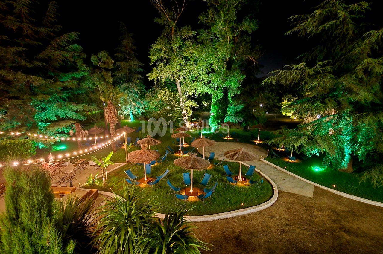 Jardin de nuit avec pelouse, chaises longues bleues sous parasols et arbres illuminés en vert.