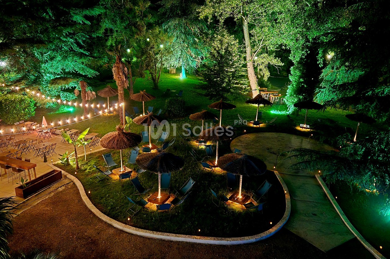 Espace événementiel nocturne avec ilots de sièges sous parasols en paille, éclairage vert et blanc, allées pavées, pelouses et arbres.