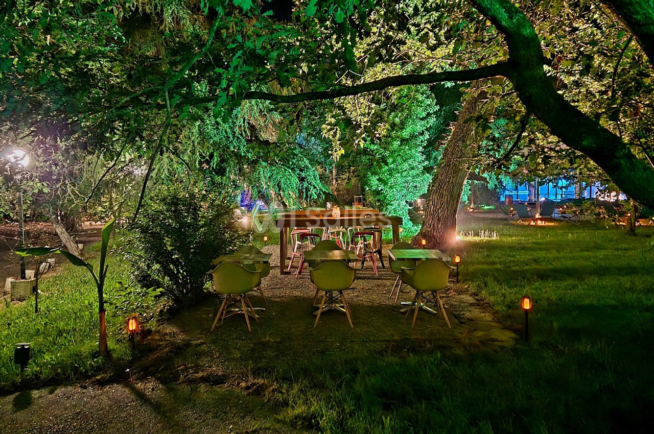 Jardin arboré de nuit avec tables dressées sous les arbres, chaises vertes, éclairage led vert et lanternes.