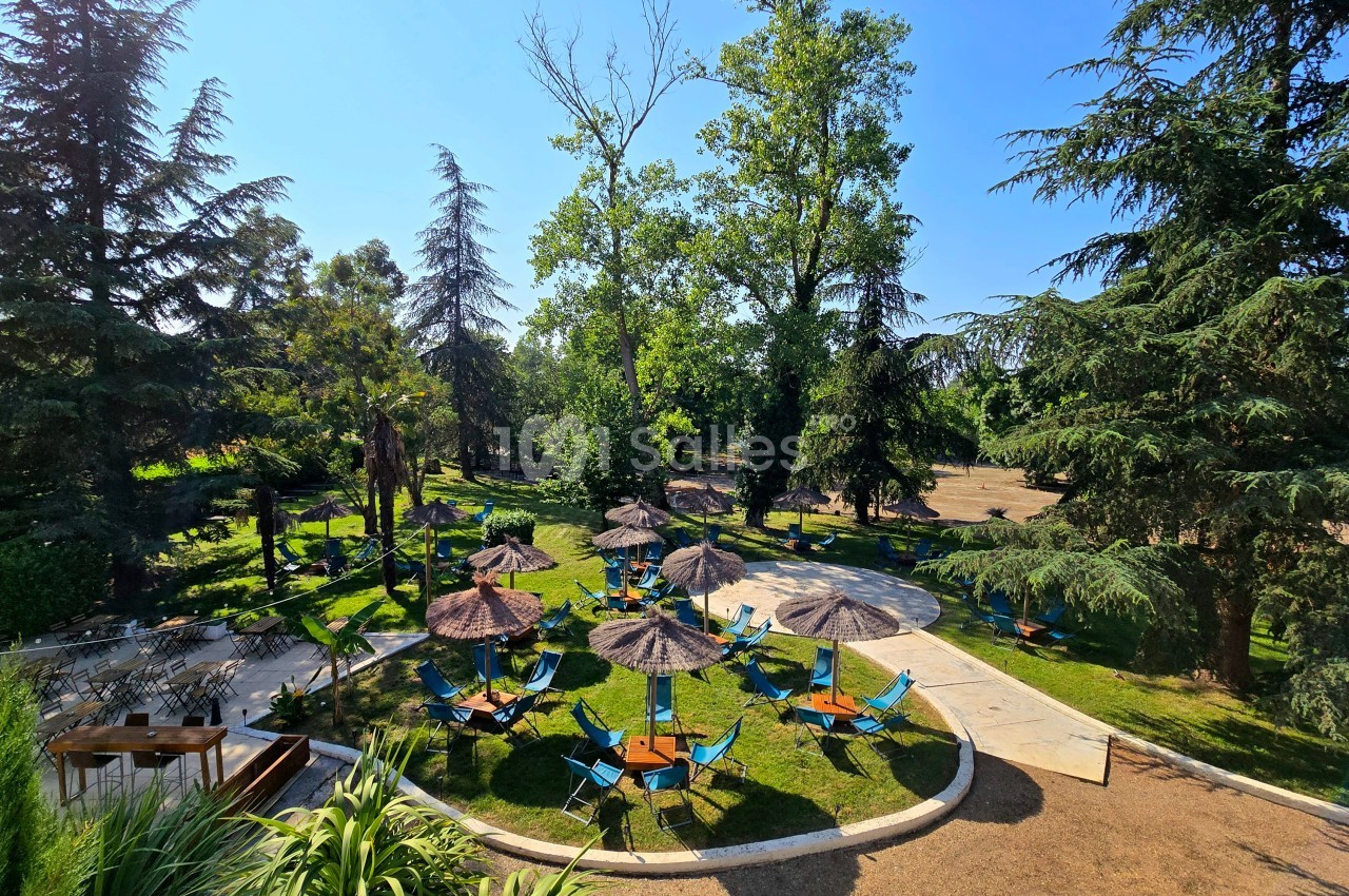 Parc arboré avec pelouse, transats bleus sous parasols et terrasse pavée en arrière-plan.