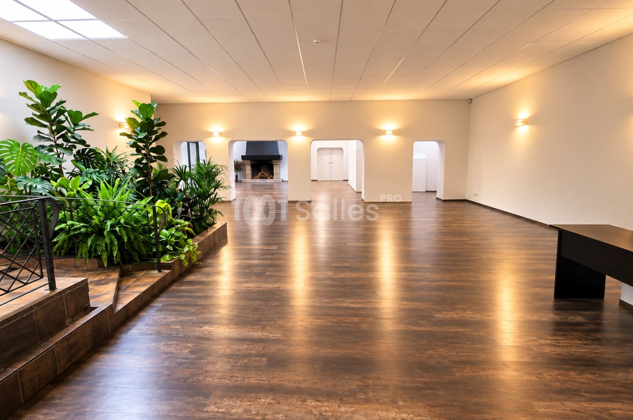 Salle vide avec sol en parquet foncé, plantes vertes sur le côté gauche et éclairage encastré chaud au plafond.