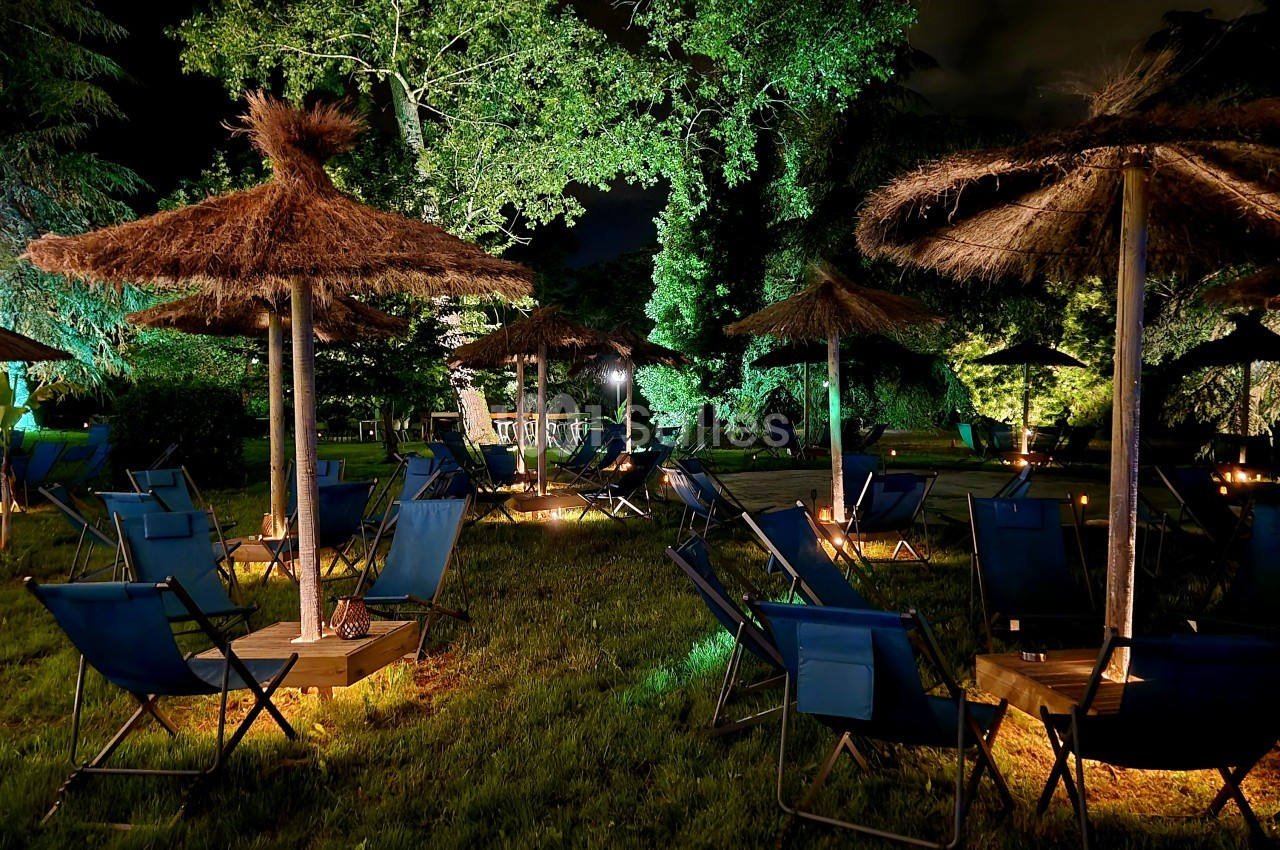 Espace extérieur de nuit avec chaises longues bleues sous parasols paillés et arbres éclairés.
