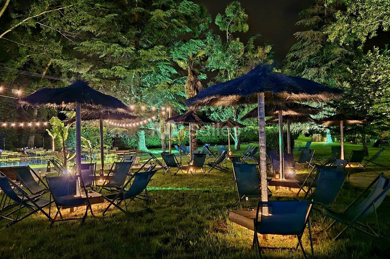 Espace extérieur de nuit aménagé avec chaises longues, parasols de paille et guirlandes lumineuses sous les arbres.