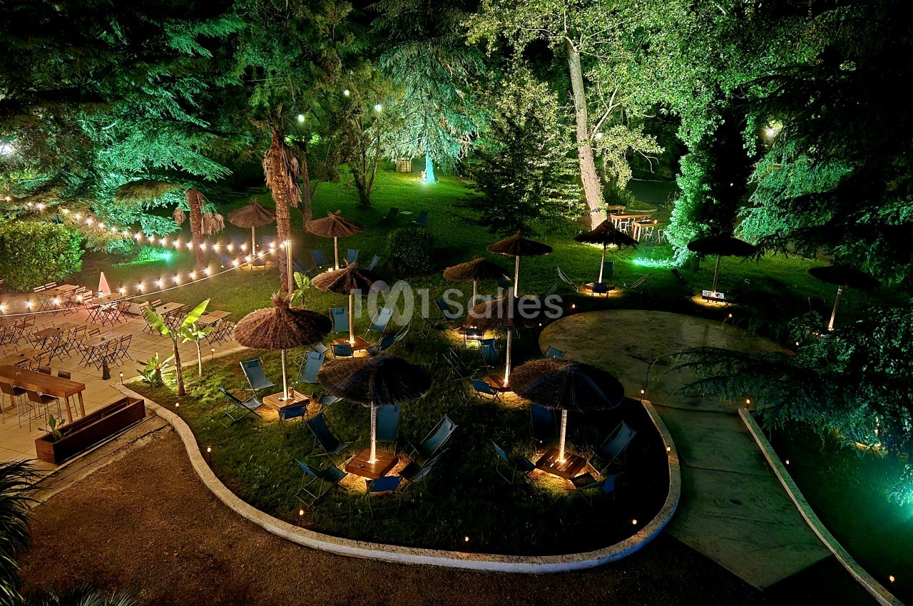Jardin nocturne aménagé avec chaises longues sous parasols, guirlandes lumineuses et arbres éclairés en vert.