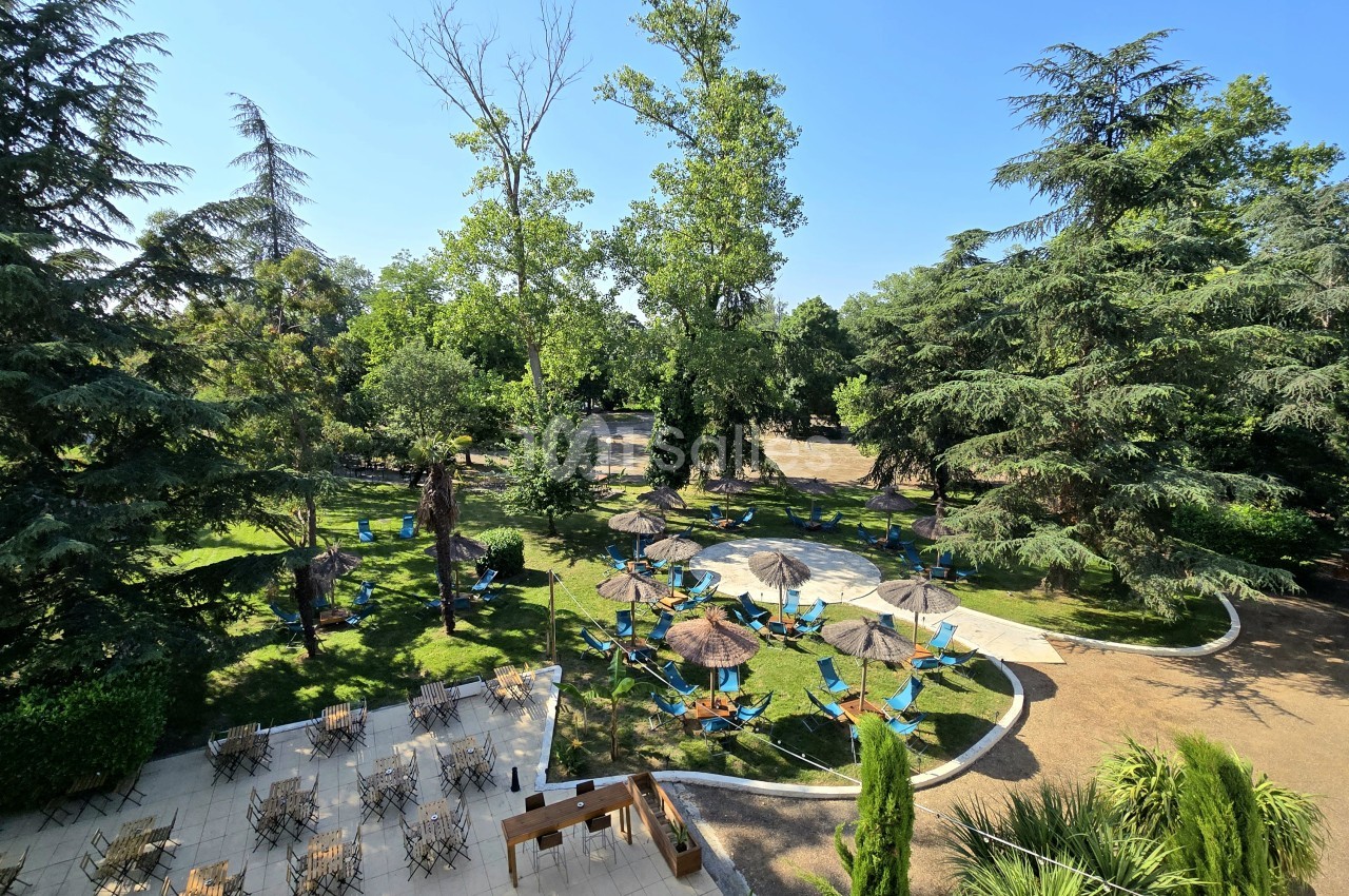 Parc boisé avec terrasse dallée, chaises pliantes, parasols paille et chemins sinueux en terre.