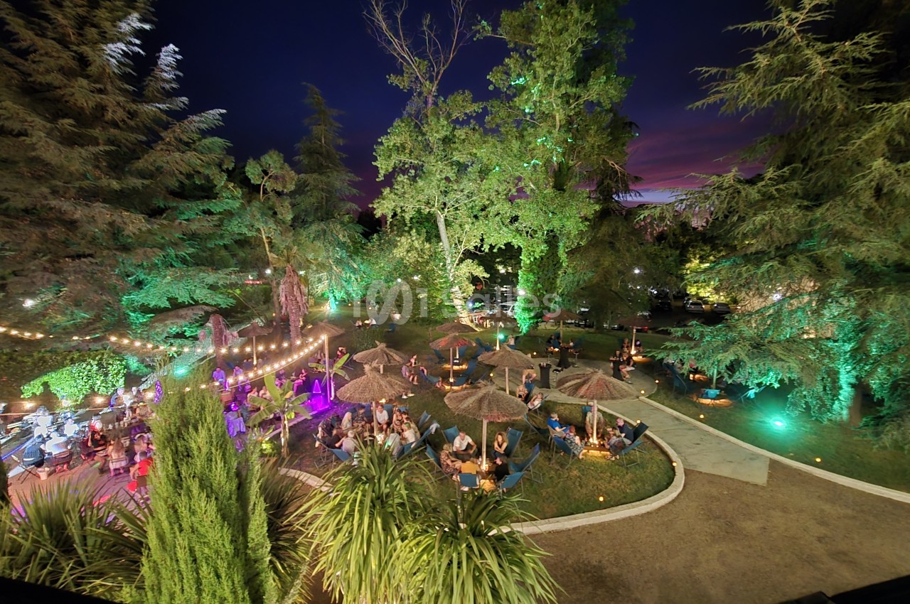 Jardin nocturne illuminé avec parasols, convives attablés sous les arbres, scène musicale à gauche et éclairage vert des végétaux.