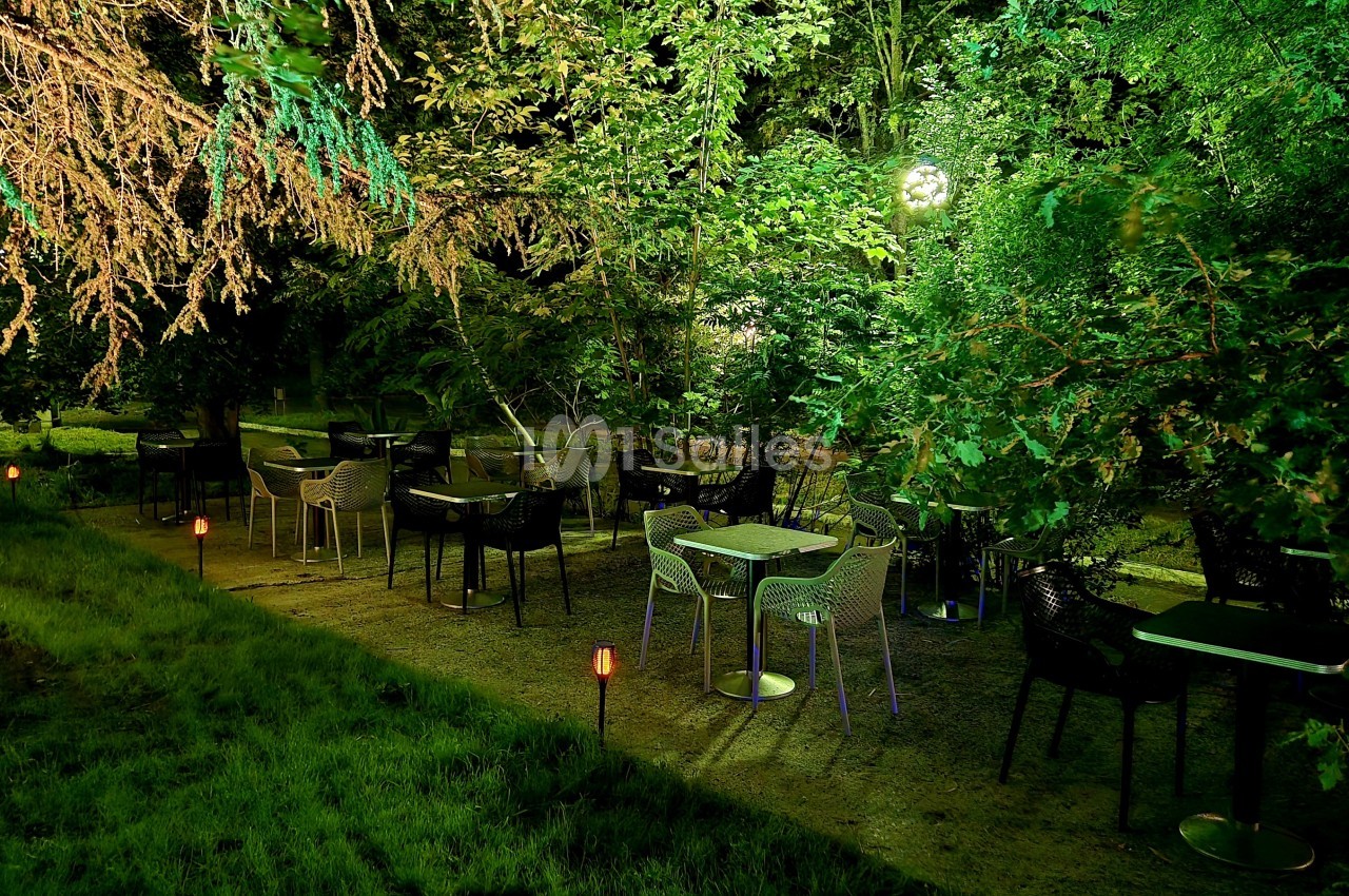 Terrasse extérieure sous la canopée avec tables et chaises noires, éclairage indirect au sol.
